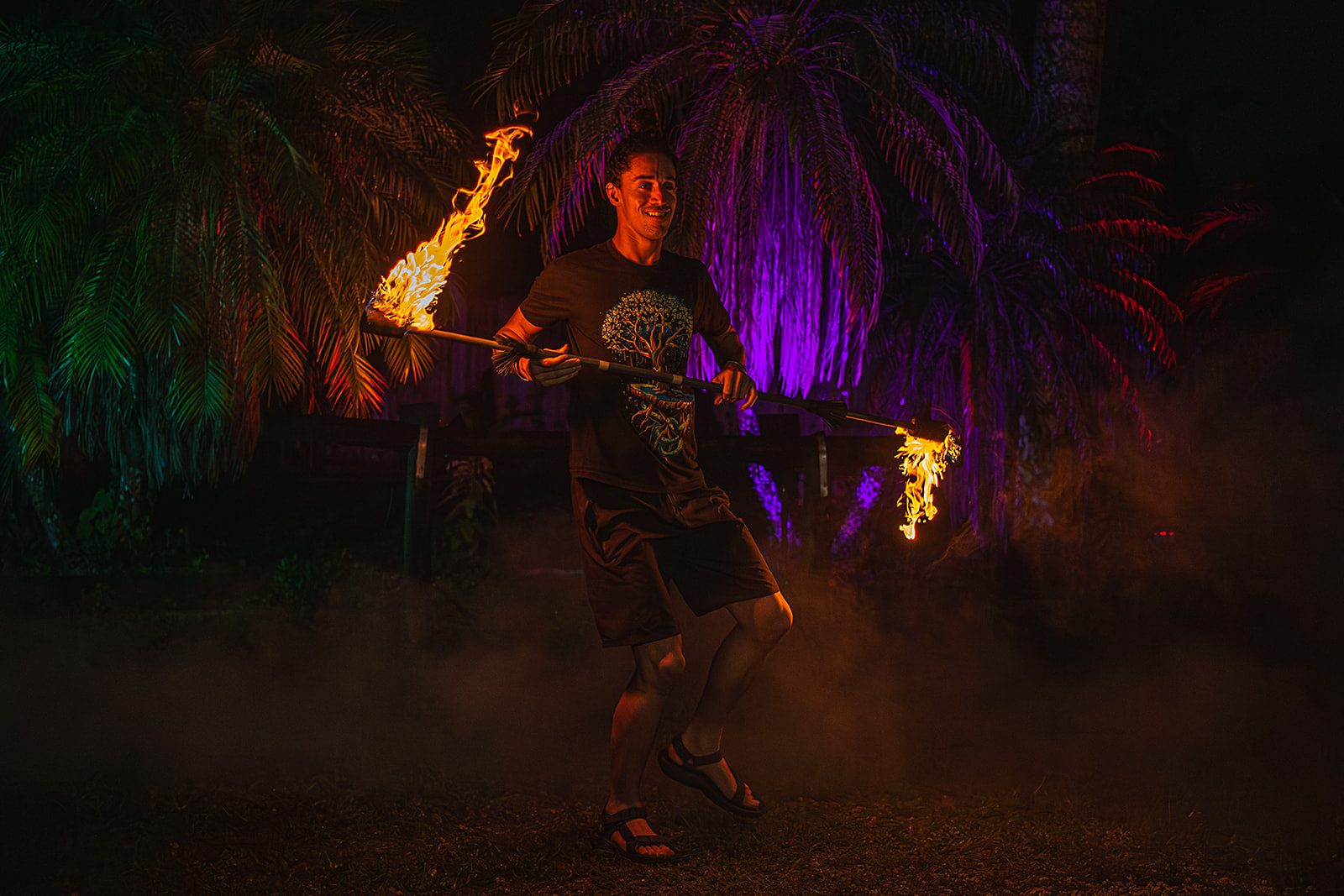 Jake walking with a lit fire staff across his body, grinning, under purple-lit palms