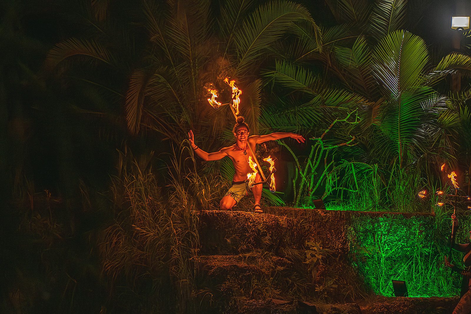 Jake kneeling on stone steps in a green-lit jungle, arms open, a lit fire staff balanced across his shoulders