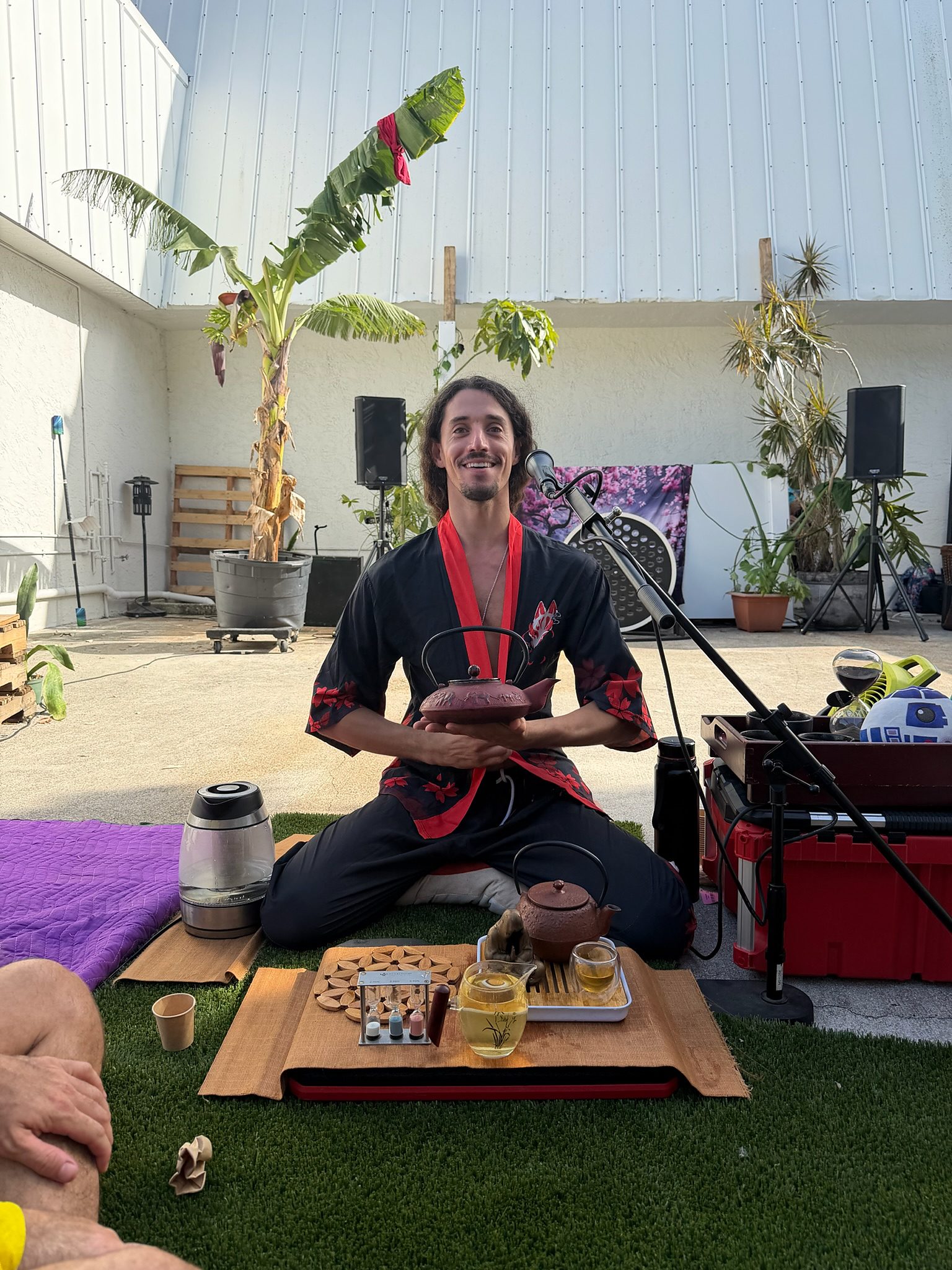 Jake seated on turf in a black and red kimono, holding a clay teapot, a microphone beside him